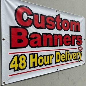 May include: White banner with the words "Custom Banners" in red and black lettering. Below, the text "48 Hour Delivery" is in yellow and black. The banner is hanging on a gray wall.