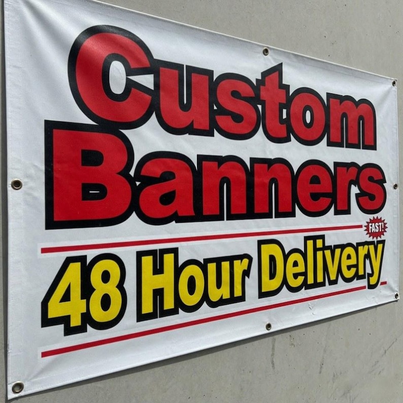 May include: White banner with the words "Custom Banners" in red and black lettering. Below, the text "48 Hour Delivery" is in yellow and black. The banner is hanging on a gray wall.