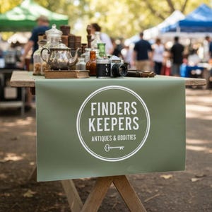 May include: A vintage market display featuring a green table runner with the text "FINDERS KEEPERS ANTIQUES & ODDITIES". The table holds a silver teapot, antique camera, and various glass bottles.