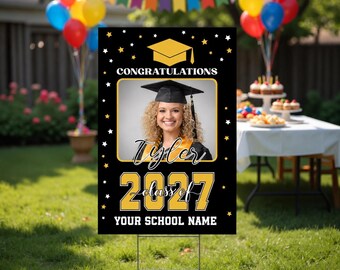 May include: A black rectangular graduation yard sign featuring a yellow mortarboard icon, the text CONGRATULATIONS, a central photo frame, the name Tyler, the year 2027, and the text YOUR SCHOOL NAME, decorated with small white and yellow stars.