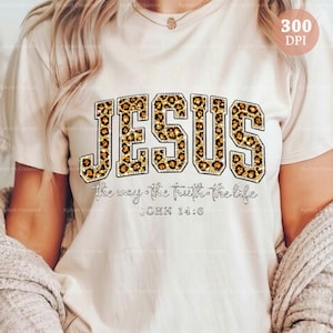 May include: A cream-coloured t-shirt displaying the word "JESUS" in large leopard print letters with a rhinestone outline. Beneath, the text reads "The way, the truth, the life" and "JOHN 14:6" in a script font.