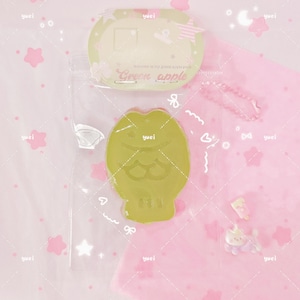 May include: A green apple-shaped item with the text "Green apple" and a fish design. The item is packaged in a clear plastic bag. The background is pink with star and flower shapes.