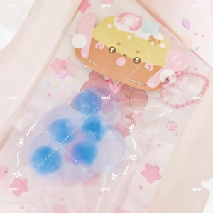 May include: A clear plastic bag containing a cartoon cupcake-shaped charm and a paw-shaped ice pack with blue gel beads. The cupcake charm has a yellow top, brown base, and pink accents. The bag has pink and white decorative elements.