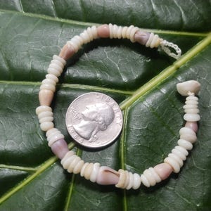 May include: A handmade seashell bracelet with a quarter coin next to it. The bracelet is made of small, white and pink seashells strung together. The quarter is silver and shows the profile of a person. The bracelet is on a large green leaf.
