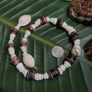 May include: A white and brown shell necklace with a gold clasp. The necklace is made of various sized shells and is laid out on a green leaf.