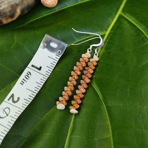 May include: A pair of handmade earrings with a beaded design. The earrings are made of small, light brown beads and white beads, strung together to create a dangling effect. The earrings are approximately 2 inches long, with silver-colored ear wires. The earrings are displayed on a green leaf.