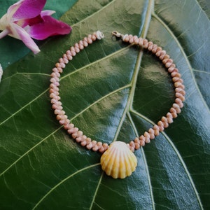 May include: A pink and white shell necklace with a large, yellow and white shell pendant. The necklace is lying on a green leaf.
