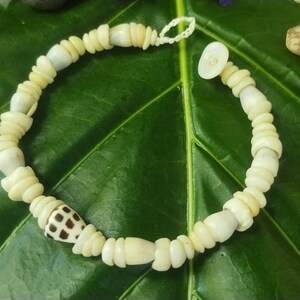 May include: A cream-colored seashell bracelet with a variety of shell shapes and sizes. The bracelet features a mix of smooth and textured shells, including one with a brown and white pattern. The bracelet is displayed on a large green leaf.