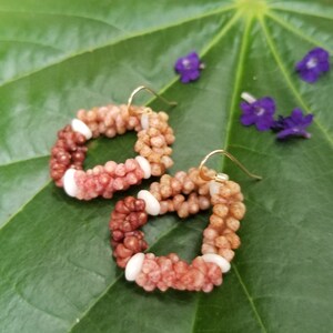 May include: A pair of hoop earrings made of small, colorful, shell beads. The earrings are a gradient of pink, red, and brown colors. The earrings are on a green leaf background.