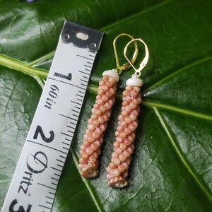 May include: A pair of handmade earrings with a spiral design, featuring small, light-brown beads. The earrings have gold-colored hooks and measure approximately 2 inches in length. The earrings are displayed on a green leaf next to a measuring tape.