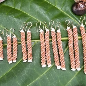 May include: A collection of handmade earrings featuring a spiral design made of small, light brown beads. The earrings have silver-colored hooks and white accents at the top and bottom. They are displayed on a large green leaf.