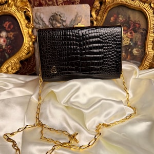 Peut inclure: Une pochette rectangulaire noire avec une bandouli&egrave;re en cha&icirc;ne dor&eacute;e. La pochette a une texture peau de crocodile et un fermoir dor&eacute;. La cha&icirc;ne est dor&eacute;e et le sac repose sur une surface en satin blanc. Un collier de perles est au premier plan.