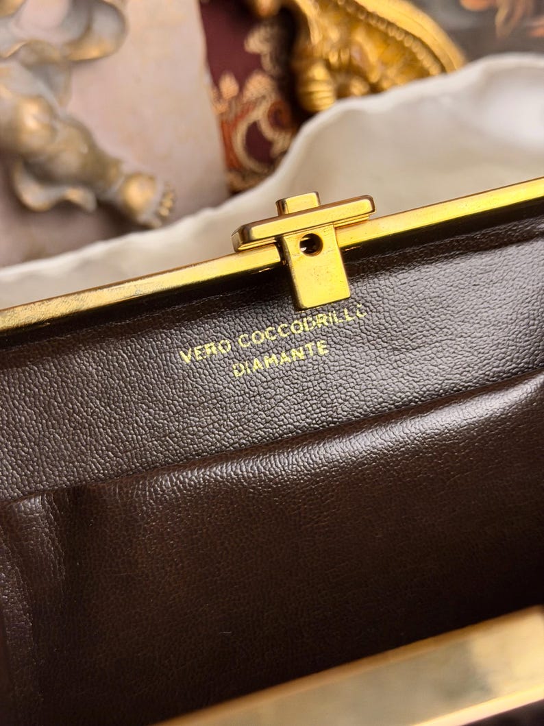 Peut inclure: Gros plan sur un sac &agrave; main en cuir marron avec un fermoir dor&eacute;. Le texte "VERO COCCODRILLO DIAMANTE" est estamp&eacute; en or sur le cuir. Le fermoir est rectangulaire avec un petit trou au centre.