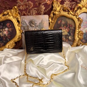 Peut inclure: Pochette rectangulaire noire avec une texture peau de crocodile, fermoir dor&eacute; et cha&icirc;ne. Le sac comporte un petit embl&egrave;me dor&eacute;. La pochette est pos&eacute;e sur une surface en satin blanc, avec des &oelig;uvres d'art encadr&eacute;es en or en arri&egrave;re-plan.