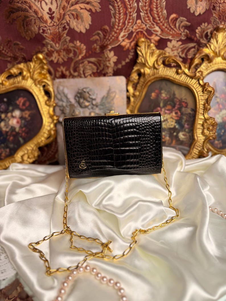 Peut inclure: Pochette rectangulaire noire avec une texture peau de crocodile, fermoir dor&eacute; et cha&icirc;ne. Le sac comporte un petit embl&egrave;me dor&eacute;. La pochette est pos&eacute;e sur une surface en satin blanc, avec des &oelig;uvres d'art encadr&eacute;es en or en arri&egrave;re-plan.