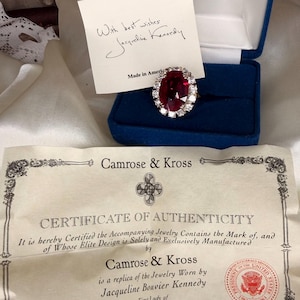 Peut inclure: Une bague rouge rubis entourée de pierres blanches, présentée dans un écrin de velours bleu. Une note manuscrite de Jacqueline Kennedy et un certificat d'authenticité de Camrose & Kross sont visibles.