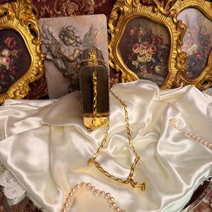 Peut inclure: Un sac &agrave; main rectangulaire vintage avec une cha&icirc;ne dor&eacute;e. Le sac, de couleur sombre, pr&eacute;sente des d&eacute;tails dor&eacute;s et un fermoir. Il repose sur un drap&eacute; de satin blanc, avec un collier de perles et des cadres dor&eacute;s orn&eacute;s en arri&egrave;re-plan.