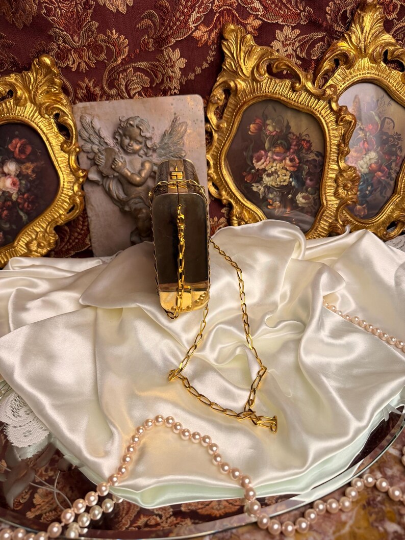 Peut inclure: Un sac &agrave; main rectangulaire vintage avec une cha&icirc;ne dor&eacute;e. Le sac, de couleur sombre, pr&eacute;sente des d&eacute;tails dor&eacute;s et un fermoir. Il repose sur un drap&eacute; de satin blanc, avec un collier de perles et des cadres dor&eacute;s orn&eacute;s en arri&egrave;re-plan.