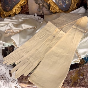 May include: A pair of long, pale yellow gloves, possibly vintage, are displayed. The gloves are made of a soft material and have a buttoned closure. A string of pearls and lace are in the background.