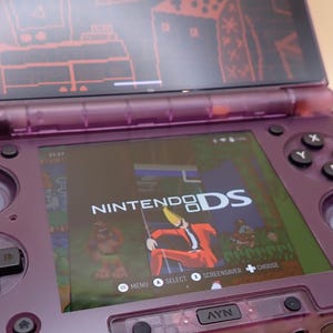 May include: A purple Nintendo DS handheld game console, open to display the screen. The screen shows a game with the text "NINTENDO DS". The console has a black directional pad, buttons, and a stylus. The top screen has a red graphic.