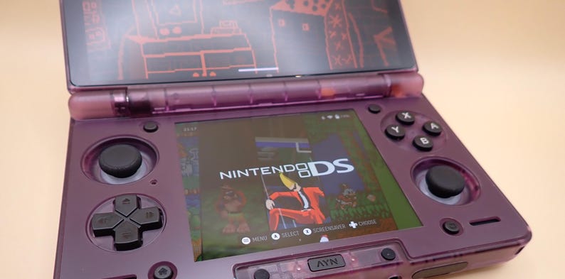 May include: A purple Nintendo DS handheld game console, open to display the screen. The screen shows a game with the text "NINTENDO DS". The console has a black directional pad, buttons, and a stylus. The top screen has a red graphic.