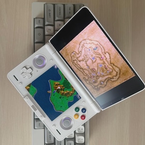May include: A white handheld gaming console with two screens, one displaying a map and the other a game scene. It rests on a gray mechanical keyboard. The console has colored buttons and a directional pad.