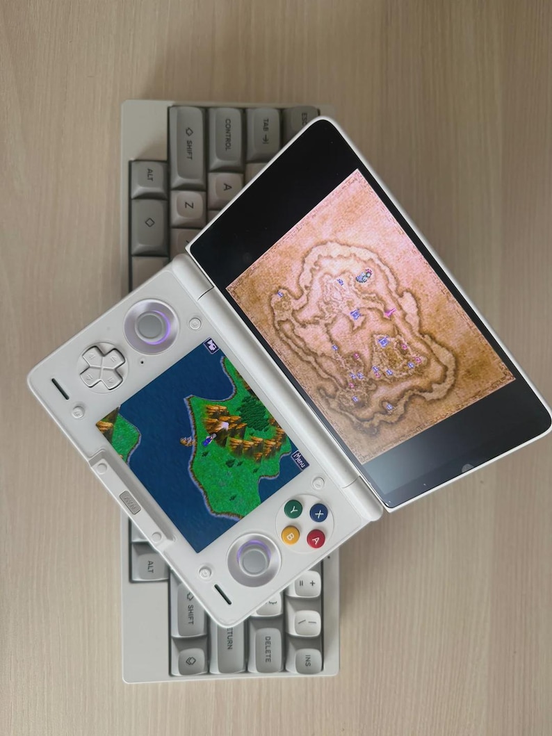 May include: A white handheld gaming console with two screens, one displaying a map and the other a game scene. It rests on a gray mechanical keyboard. The console has colored buttons and a directional pad.