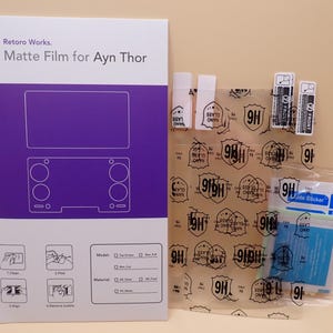 May include: A product package labeled "Retoro Works. Matte Film for Ayn Thor" with a purple and white design. The package includes a screen protector, cleaning wipes, and application tools. The screen protector has a 9H rating.