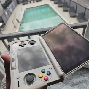 May include: A light gray handheld gaming device with a connected tablet. The device has a directional pad, a gray joystick, and colored action buttons. The tablet screen reflects the sky. The device is held in a hand.