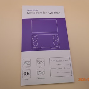 May include: A white and purple matte film sheet for the Ayn Thor handheld gaming device. The sheet features a diagram of the device and instructions for application. The text "Retoro Works. Matte Film for Ayn Thor" is visible.