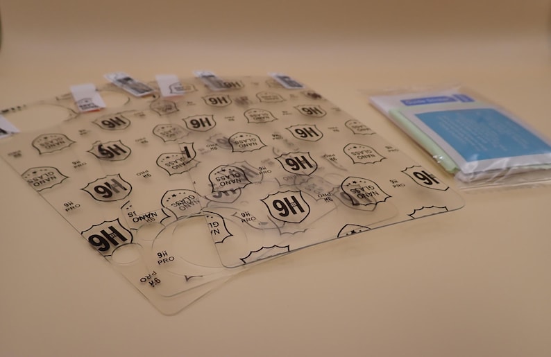 May include: A collection of clear screen protectors with the text "9H Nano Glass Pro" printed on them. The protectors are arranged on a light surface, with a small packet of cleaning supplies visible in the background.