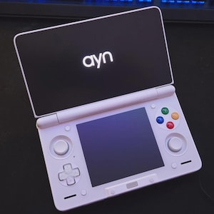 May include: A white handheld gaming console with a black top screen featuring the word "ayn" in white. The console has a lower screen, a directional pad, colored buttons, and control sticks. The device is open and ready for use.