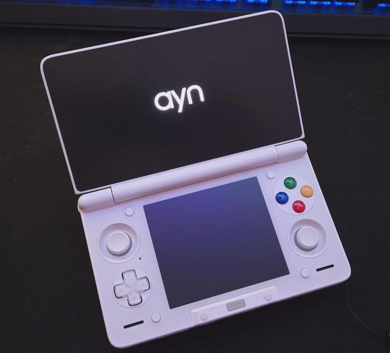 May include: A white handheld gaming console with a black top screen featuring the word "ayn" in white. The console has a lower screen, a directional pad, colored buttons, and control sticks. The device is open and ready for use.