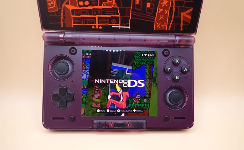 May include: A purple Nintendo DS handheld game console, open to display a game screen. The screen shows the "Nintendo DS" logo and a colorful game scene. The console has a D-pad, buttons, and a top screen with an orange pixelated design.