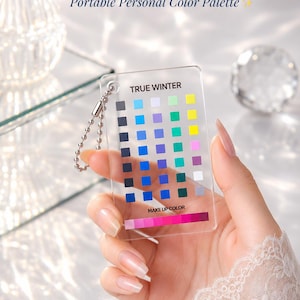 Może przedstawiać: Przezroczysta, prostokątna plastikowa karta z napisem TRUE WINTER oraz Portable Personal Color Palette, zawierająca siatkę małych kolorowych kwadratów i poziomy pasek kolorów makijażu. Do górnego rogu przymocowany jest metalowy łańcuszek kulkowy.