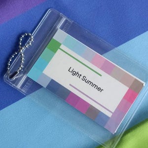 Puede incluir: Un soporte de plástico transparente con una cadena plateada contiene una tarjeta con una paleta de colores. La tarjeta dice "Light Summer" en púrpura. El fondo es una tela con rayas azules, moradas y verdes.