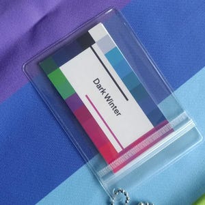 May include: A color palette card labeled "Dark Winter" inside a clear plastic sleeve with a ball chain. The card displays a range of colors, including blues, purples, and pinks. The background is a striped fabric in shades of blue, purple, and green.