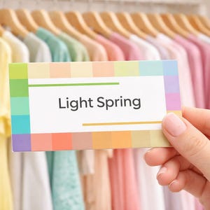 Op de afbeelding: Een kleurenpaletkaart met het label "Light Spring" wordt vastgehouden voor een kast vol pastelkleurige shirts op houten hangers. De kaart toont een reeks zachte, lenteachtige tinten.
