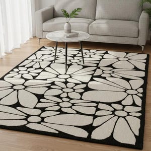 Può includere: Un tappeto rettangolare con un motivo floreale. Il tappeto presenta grandi forme di fiori bianchi stilizzati, delineati in nero, su uno sfondo nero. È posizionato su un pavimento in legno, con un divano e un tavolino sullo sfondo.