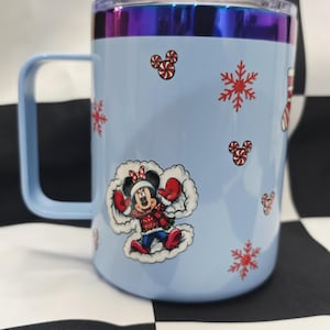 May include: A light blue travel mug with a handle and a clear lid. The mug features a Minnie Mouse graphic in a winter outfit, snowflakes, and candy cane designs. The rim has a metallic, iridescent finish.