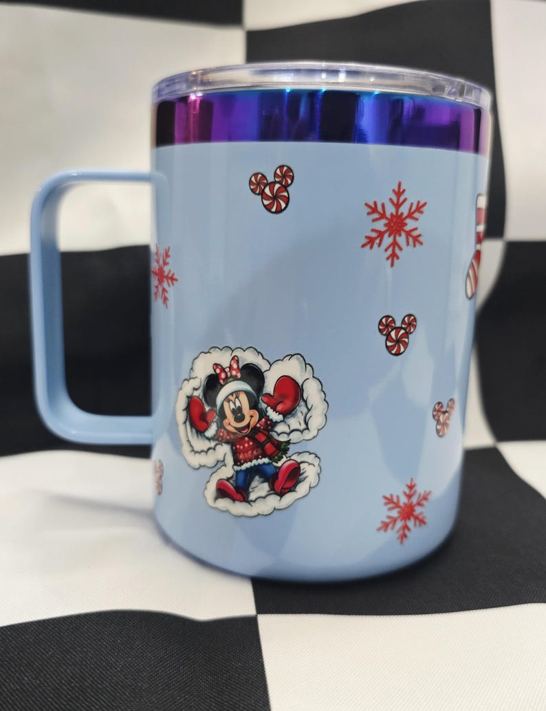 May include: A light blue travel mug with a handle and a clear lid. The mug features a Minnie Mouse graphic in a winter outfit, snowflakes, and candy cane designs. The rim has a metallic, iridescent finish.