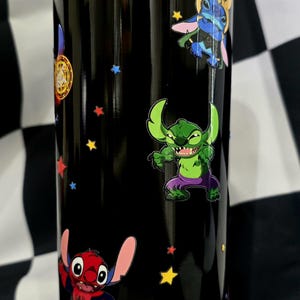 May include: Black tumbler featuring cartoon characters, including Stitch in various superhero-themed costumes, and colorful star accents. The tumbler has a metallic gold, purple, and yellow rim.