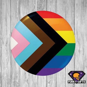 May include: A round button featuring a pride flag design. The flag has a black chevron with brown, light blue, pink, green, yellow, orange and red stripes.