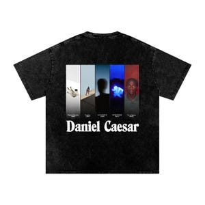 Puede incluir: Camiseta negra con un diseño gráfico blanco con el texto "Daniel Caesar". El diseño incluye paneles verticales con imágenes y texto. La camiseta tiene un aspecto vintage lavado.