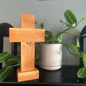 May include: A wooden cross with a natural finish, displayed on a black shelf. The cross is made of rectangular pieces of wood joined together. A potted plant with green and white striped leaves is next to the cross.