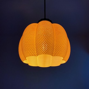 May include: A hanging, pumpkin-shaped pendant light fixture. The light has a textured, honeycomb-like surface and emits a warm, yellow glow. The fixture is suspended from a black cord against a dark blue background.