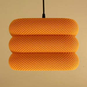 May include: A hanging, orange pendant light fixture. The light has a cylindrical shape with three stacked, rounded sections. The shade has a textured, woven appearance. A black cord suspends the light from the ceiling.
