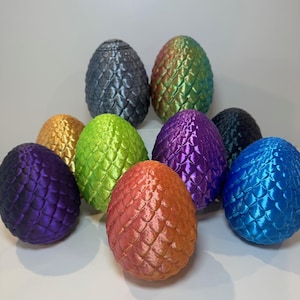 Op de afbeelding: Een verzameling van negen decoratieve draken eieren in verschillende kleuren, waaronder paars, blauw, groen, goud en oranje. Elk ei heeft een getextureerd, geschubd oppervlak en een metallic glans, waardoor een fantasiethema ontstaat.