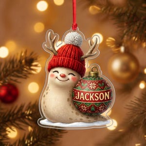 May include: A clear acrylic Christmas ornament shaped like a smiling slug wearing a red knit hat with a white pom-pom and reindeer antlers. The slug holds a red and green patterned Christmas bauble with the name JACKSON printed in white text.