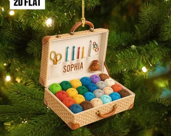 May include: A decorative Christmas ornament shaped like a suitcase, filled with colorful yarn balls. The suitcase has a handle and is decorated with crochet hooks, scissors, and the name "SOPHIA". The ornament is hanging on a Christmas tree.